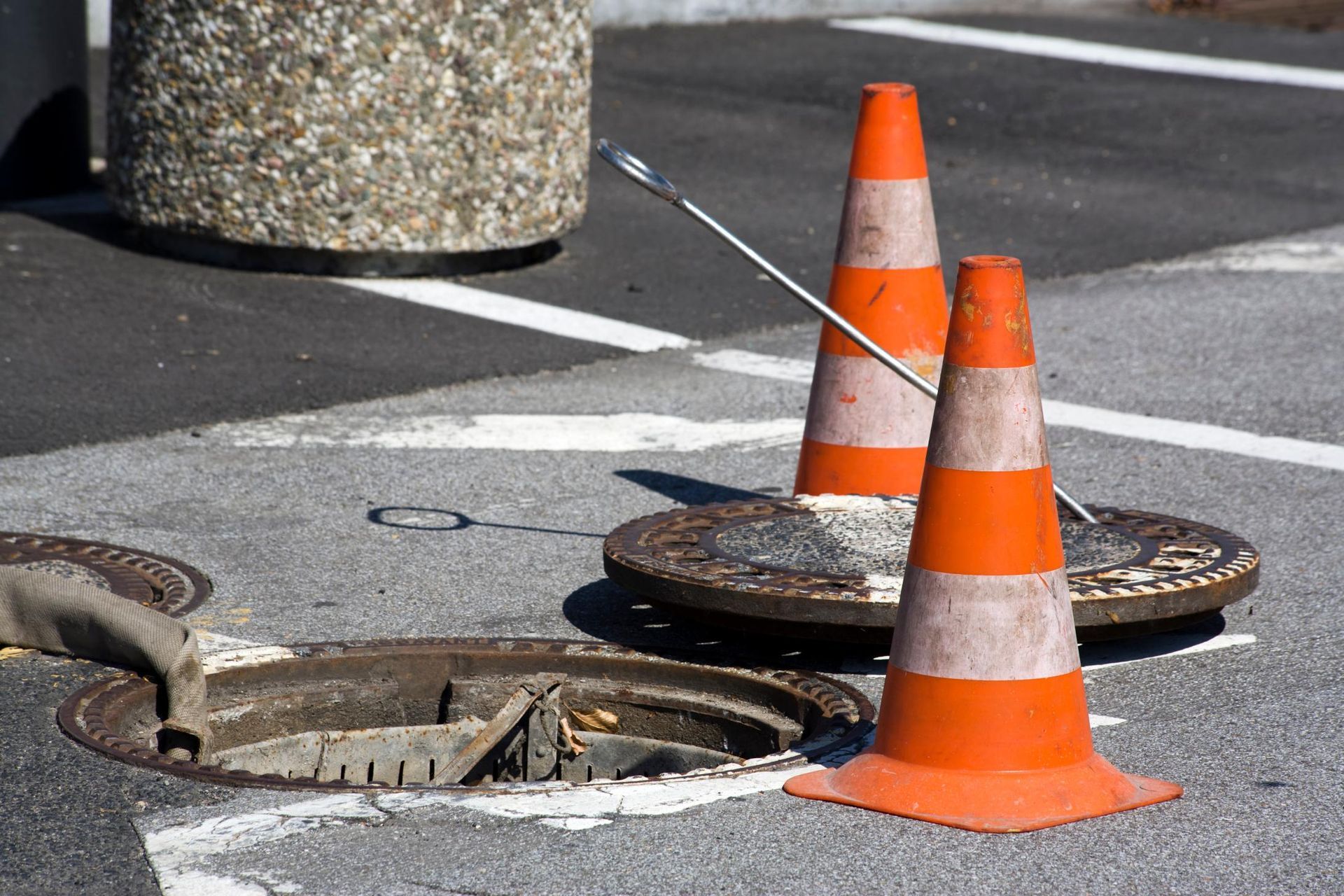 Offener Kanaldeckel auf der Straße, zwei orangefarbene Verkehrskegel und eine Metallstange warnen vor Bauarbeiten.