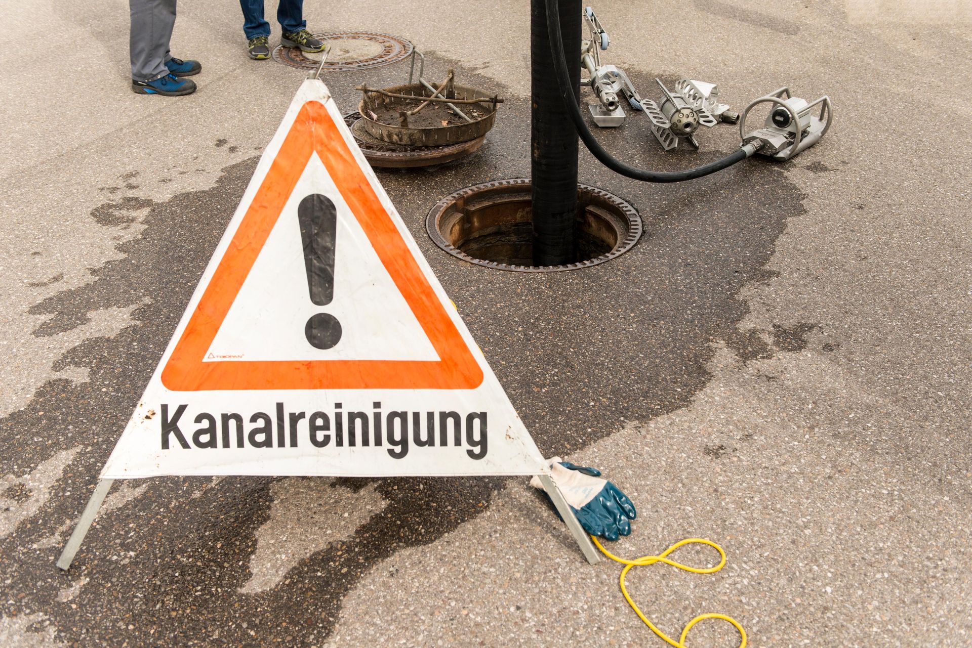 Warnschild in der Nähe eines offenen Kanaldeckels; Text „Kanalreinigung“; Kanalreinigung im Gange.