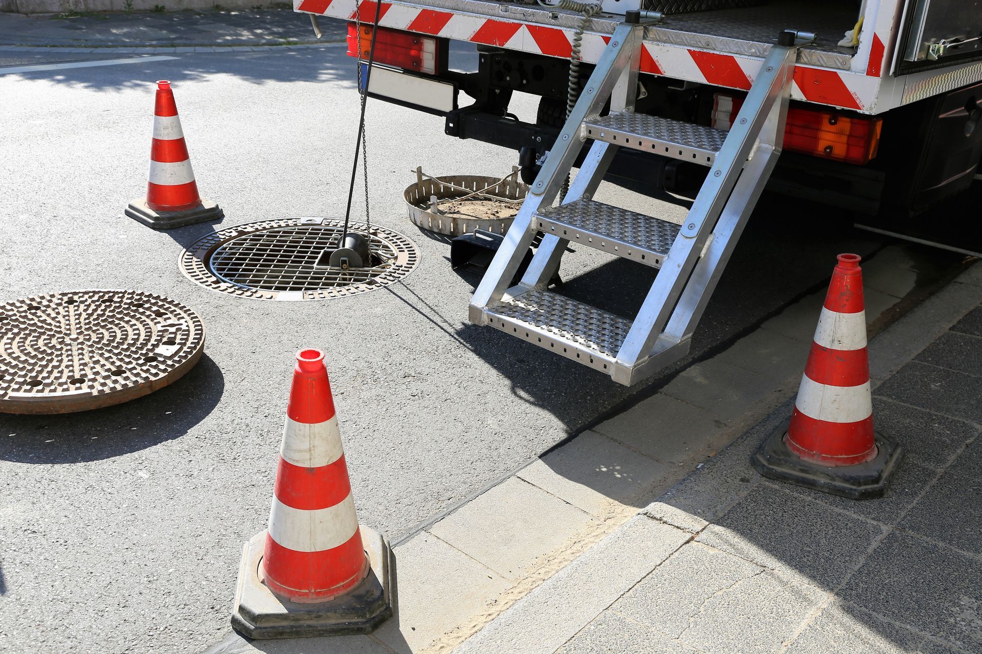 Baustelle: LKW mit Leiter, offener Kanaldeckel, Leitkegel.