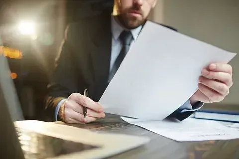 Un hombre con traje y corbata está sentado en un escritorio sosteniendo un trozo de papel y escribiendo en él.