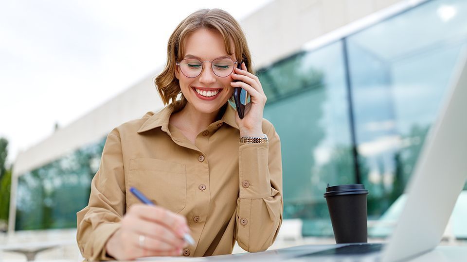 Eine Frau sitzt mit einem Laptop an einem Tisch und telefoniert mit einem Mobiltelefon.