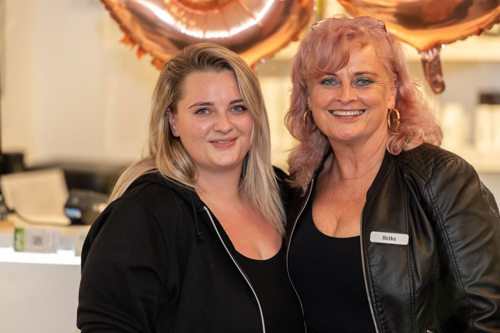 Zwei lächelnde Frauen; eine mit pinken Haaren und schwarzer Jacke. Beide tragen schwarze Oberteile. Salon-Atmosphäre.