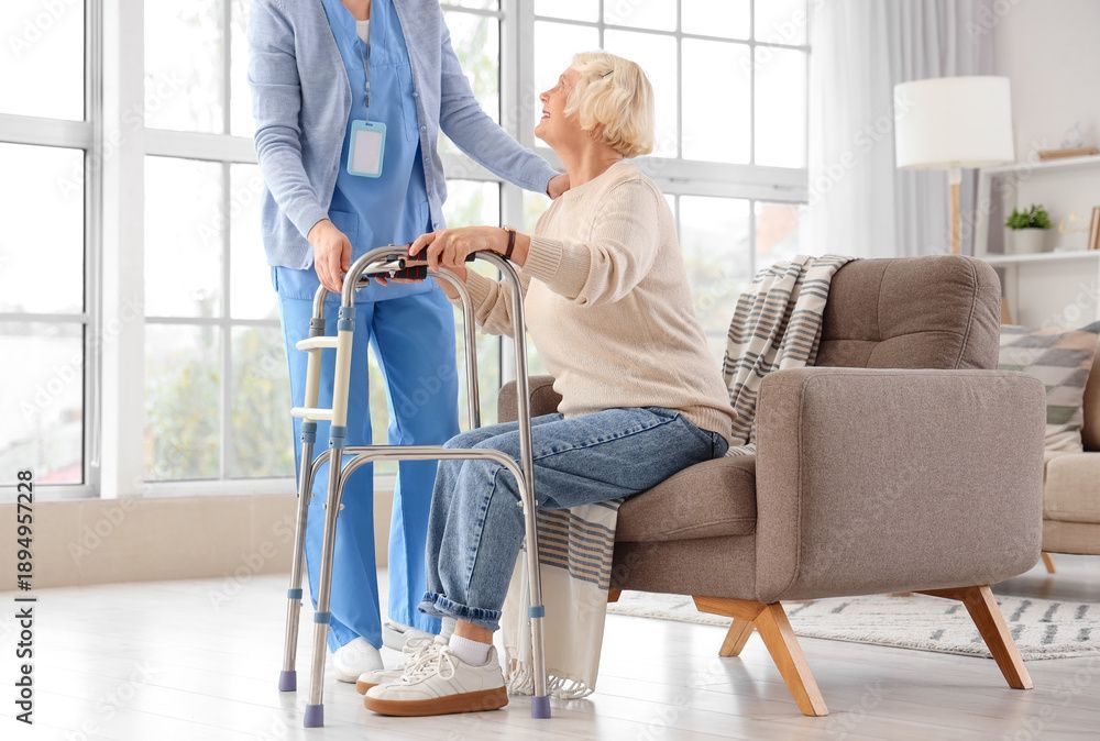 Pflegekraft hilft einem sitzenden älteren Menschen mit einem Rollator in einem hellen Wohnzimmer.