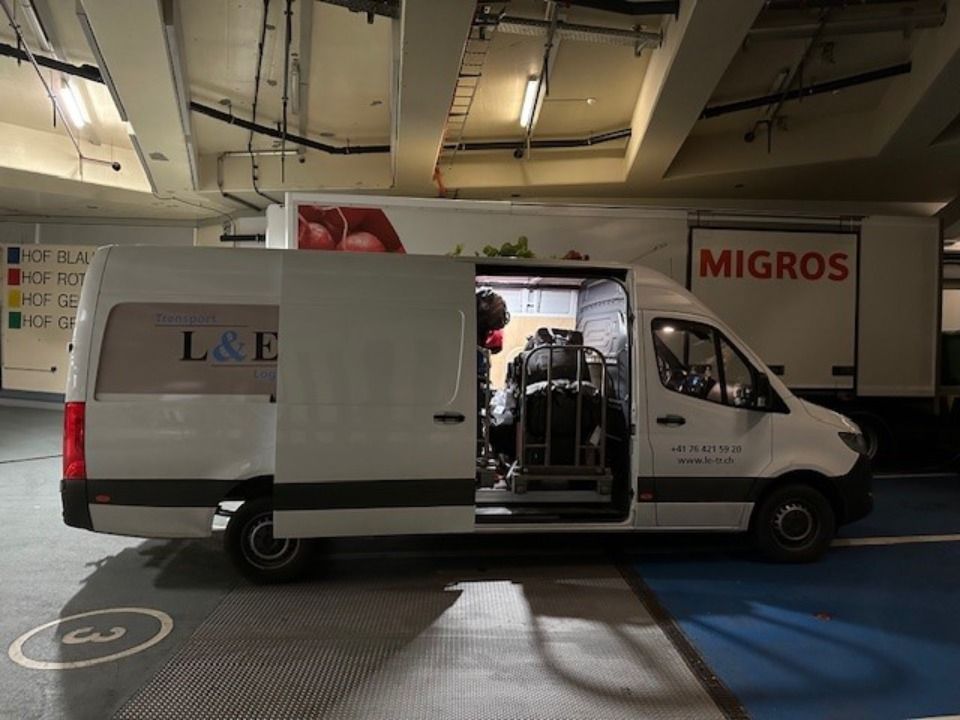 In einem Parkhaus steht ein weisser Lieferwagen neben einem grossen Migros-Lastwagen. Foto von der L&E Transport und Logistik GmbH