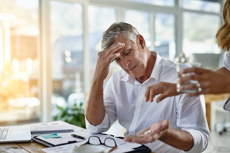 Ein Mann sitzt an einem Schreibtisch. Er hat seine Brille auf die Unterlagen vor sich gelegt und fasst sich an die Stirn. Eine Frau reicht ihm ein Glas Wasser und Tabletten. Kopfschmerzen sind ein Anzeichen für Burnout. Ein Burnout Coaching kann hier hilfreich sein. 