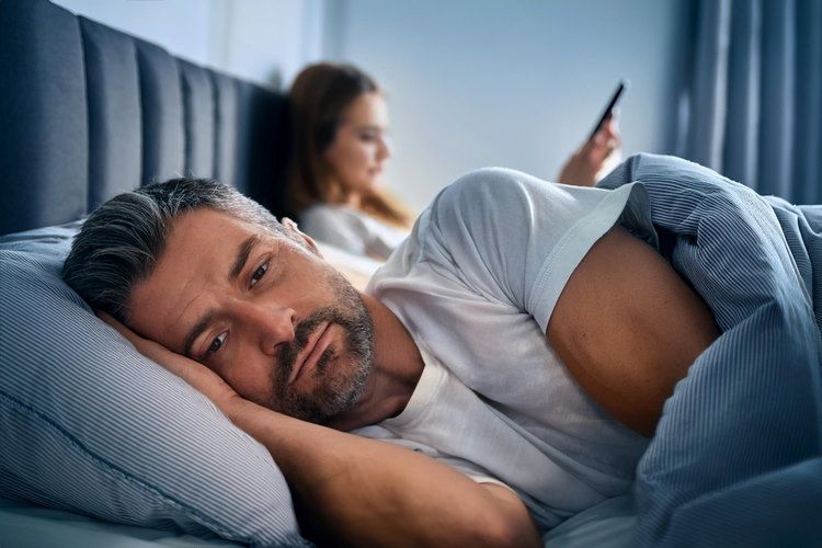 Ein Mann liegt auf der Seite im Bett. Sein Gesichtsausdruck wirkt gleichgültig, eher unzufrieden. Hinter ihm sitzt eine Frau auf ihrer Bettseite und hält ein Handy in der Hand. Die beiden wirken distanziert. Dieses Verhalten kann auf Burnout hindeuten. Ein Burnout Coach hilft Ihnen Ihre Beziehung trotz Stress wieder liebevoll zu gestalten.