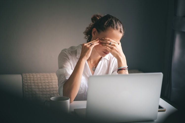 Eine Frau sitzt an ihrem Schreibtisch. Ihr geöffneter Laptop und eine Tasse stehen vor ihr. Der Raum wirkt dunkel. Die Frau hält sich erschöpft die Augen zu. Sie scheint überlastet. In solchen Fällen ist es ratsam, einen Burnout Coach aufzusuchen. 
