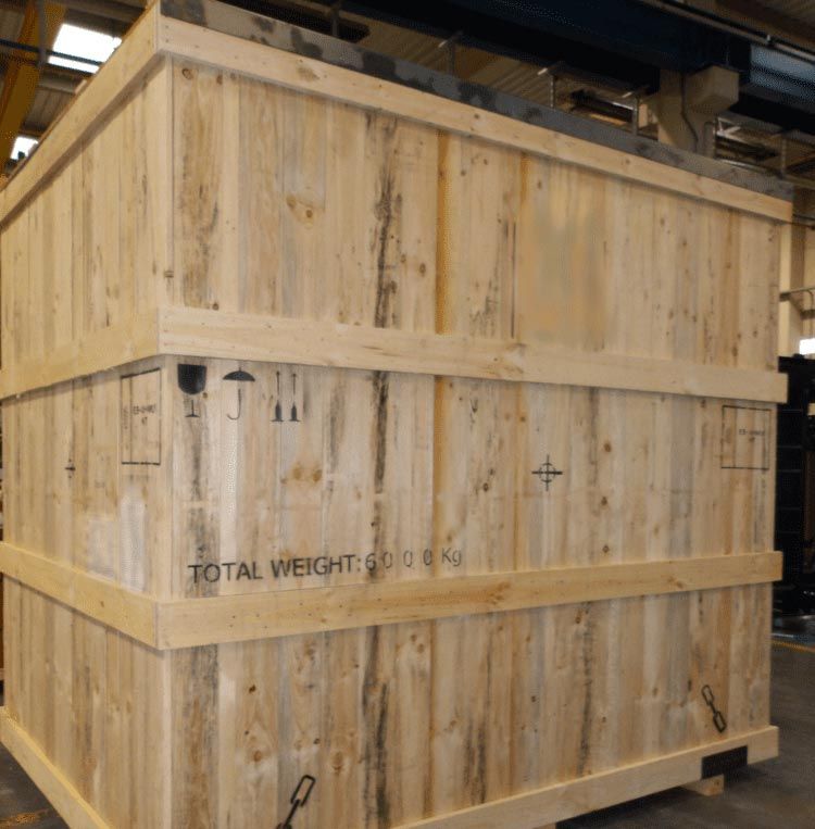 Gran caja de madera para transporte en un almacén industrial, marcada con texto y herrajes metálicos.