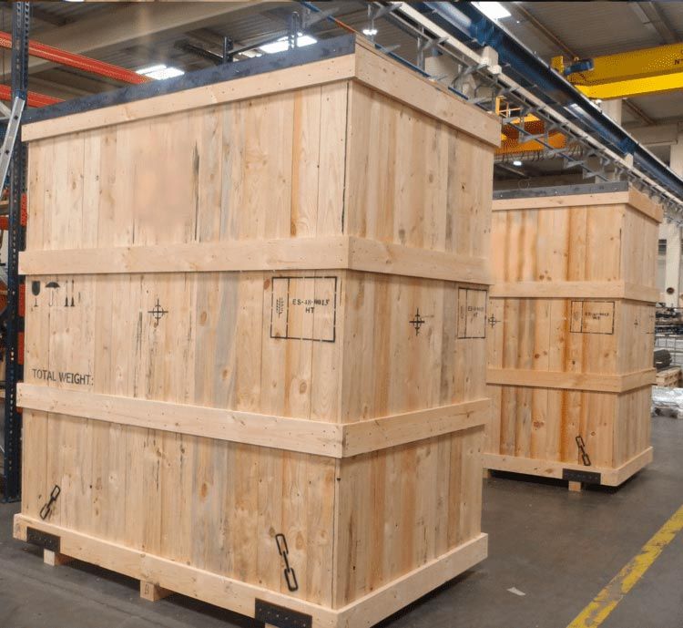 Grandes cajas de madera para transporte en un almacén industrial con grúas aéreas y suelo de hormigón.