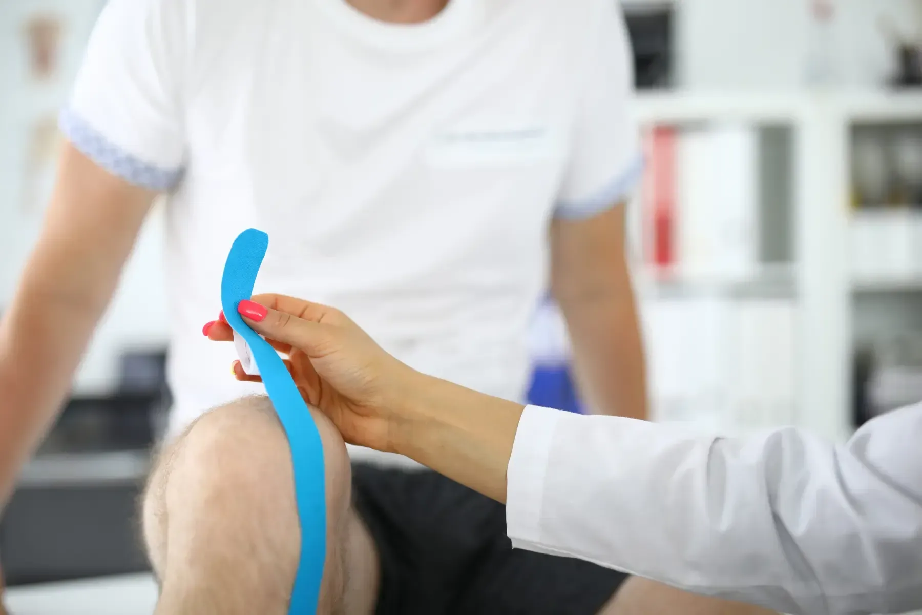 Una persona con bata médica aplica cinta kinesiológica azul en la rodilla de un paciente en un entorno clínico.