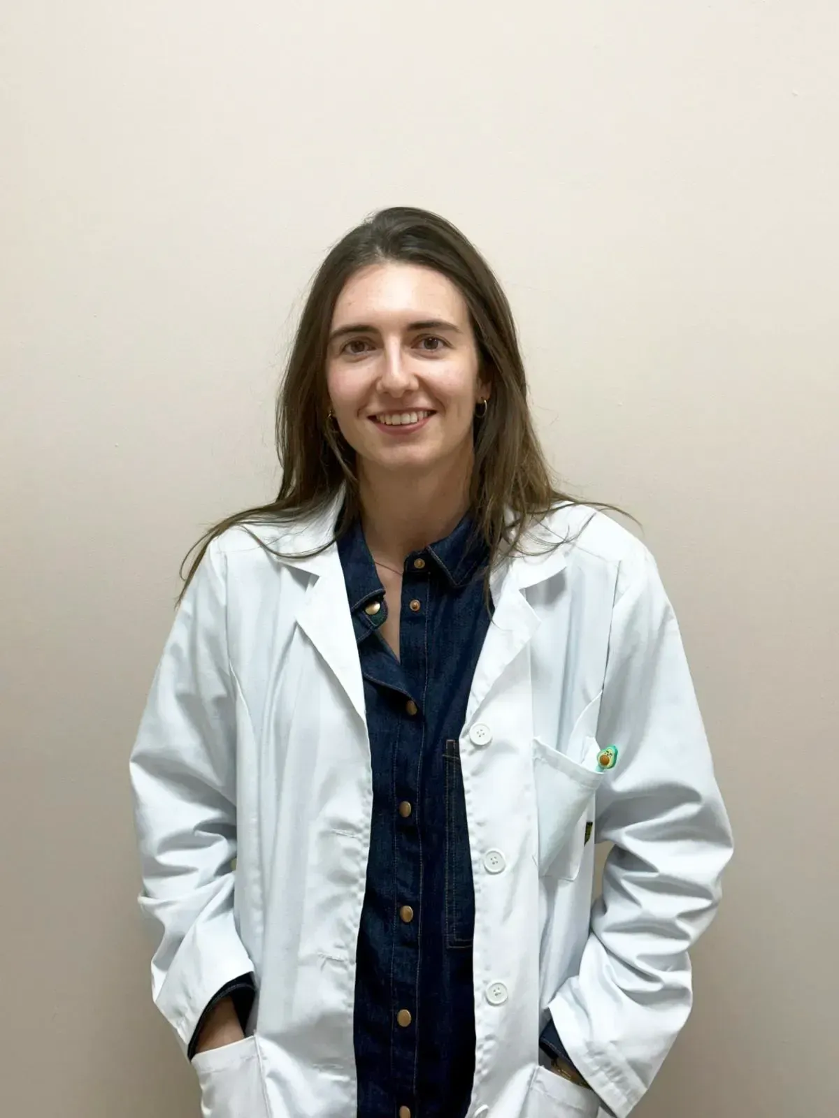 Una persona con cabello castaño largo, vestida con una bata blanca de laboratorio sobre una camisa azul oscuro abotonada.
