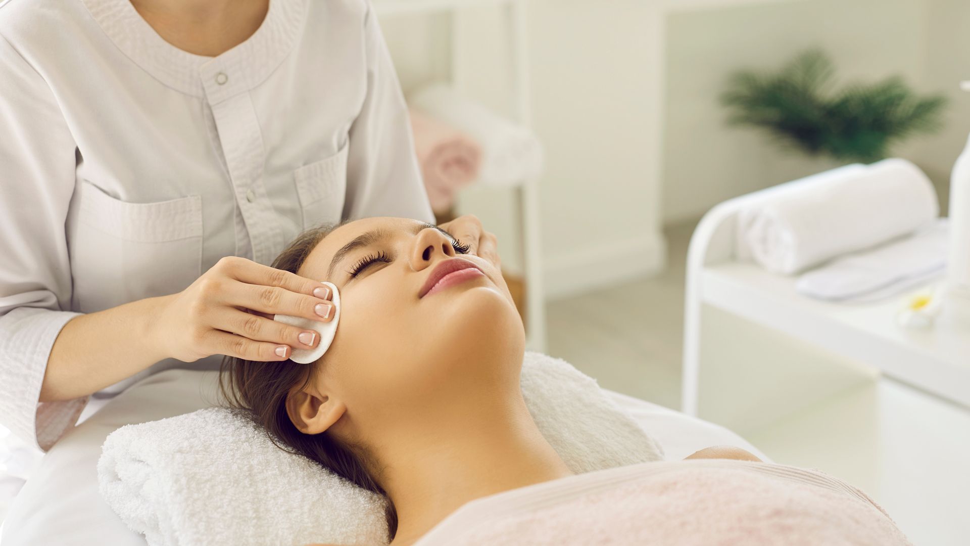 Mujer recibiendo tratamiento facial en un spa; esteticista sosteniendo un disco de algodón; interior luminoso.