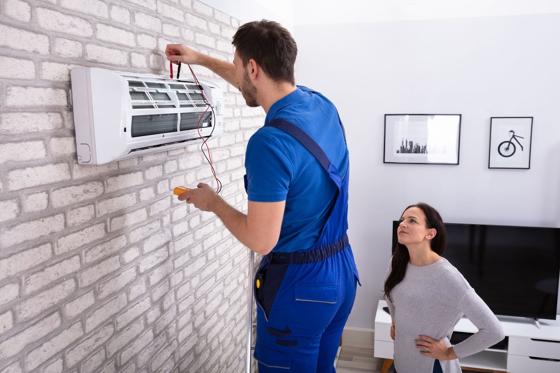 Un técnico con un mono azul repara una unidad de aire acondicionado en una pared de ladrillos, observado por una mujer con un suéter gris.
