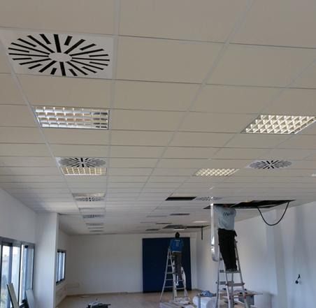 Trabajadores en escaleras instalando o reparando placas de techo en una habitación con iluminación y ventilaciones.