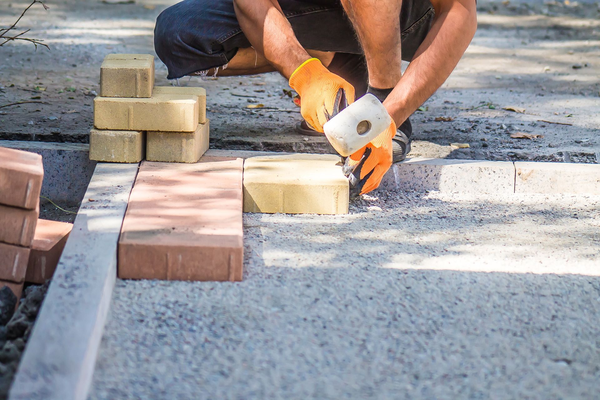 Un ouvrier pose des pavés sur une surface de gravier à l'aide d'un maillet, des blocs empilés se trouvant à proximité.