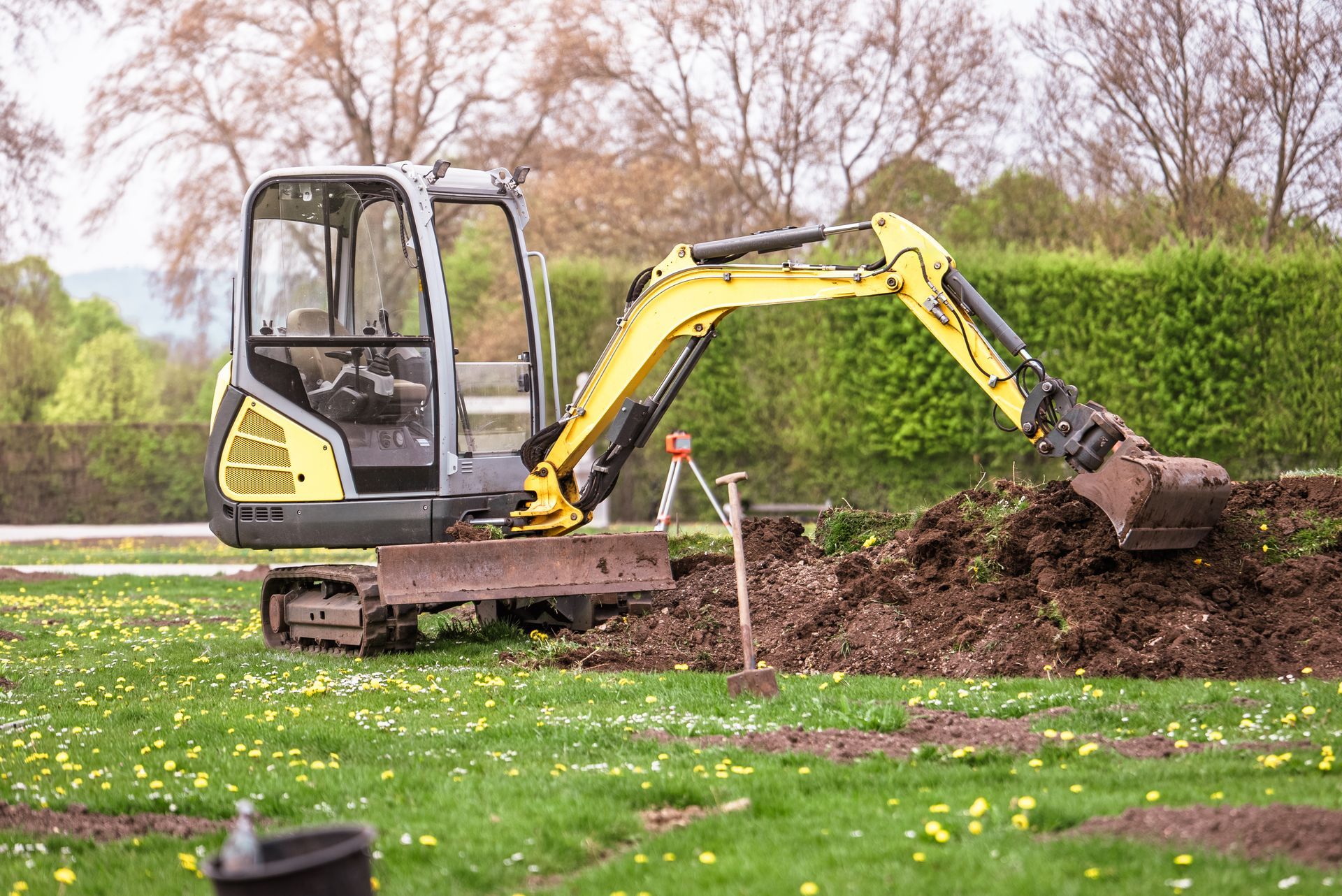 Une minipelle creuse la terre dans un jardin gazonné, près d'une haie et d'arbres dénudés.