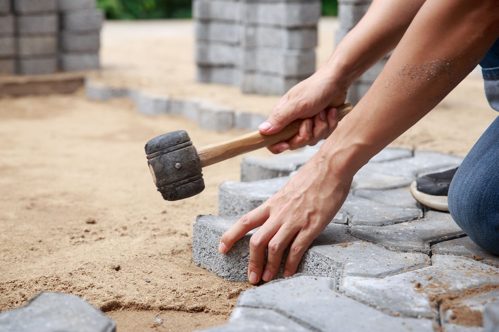 Des mains utilisent un maillet en caoutchouc pour poser des pavés gris sur du sable à l'extérieur.