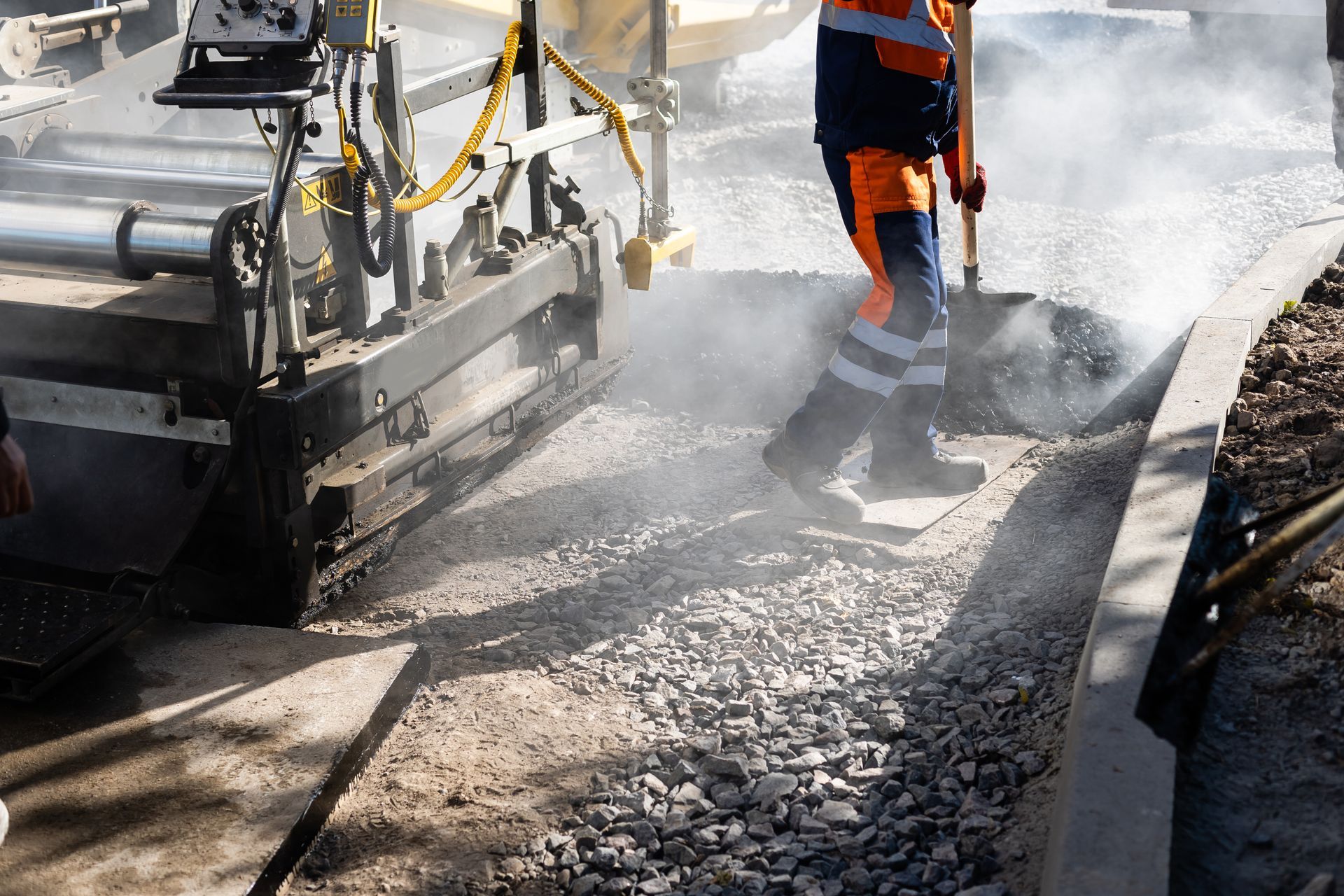 Une machine à paver la chaussée étale de l'asphalte frais à côté d'un ouvrier en tenue de sécurité orange et pantalon réfléchissant.