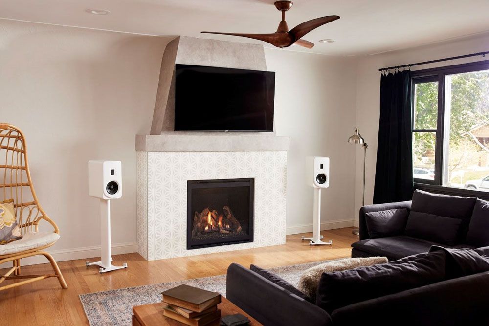 Modernes Wohnzimmer mit Kamin, Wandfernseher, dunklem Sofa, weißen Lautsprechern und großem Fenster mit Blick nach draußen