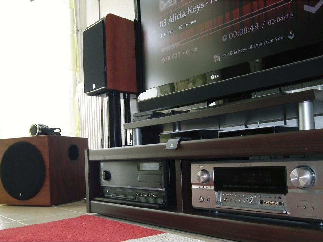 Un système audio à domicile. Des enceintes, un caisson de basses, un téléviseur et un récepteur audio vidéo sont posés sur un support en bois foncé, près d'une fenêtre.