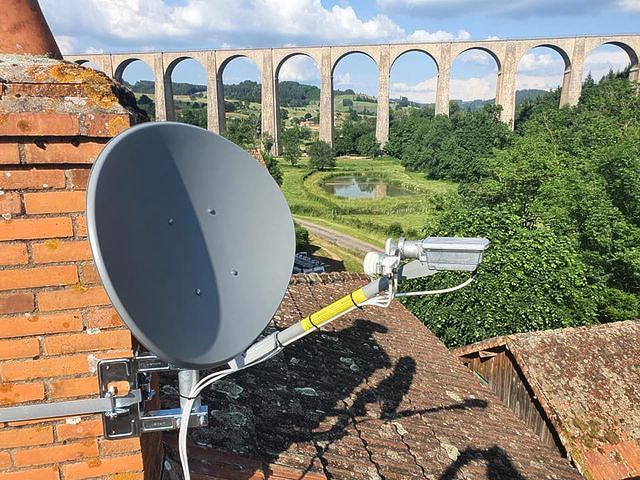 Antenne parabolique sur un toit, avec un viaduc en pierre et un paysage verdoyant en arrière-plan.