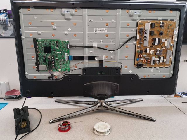 Arrière d'un téléviseur Samsung démonté, avec des cartes de circuits imprimés, des fils et un fer à souder posé sur une table.