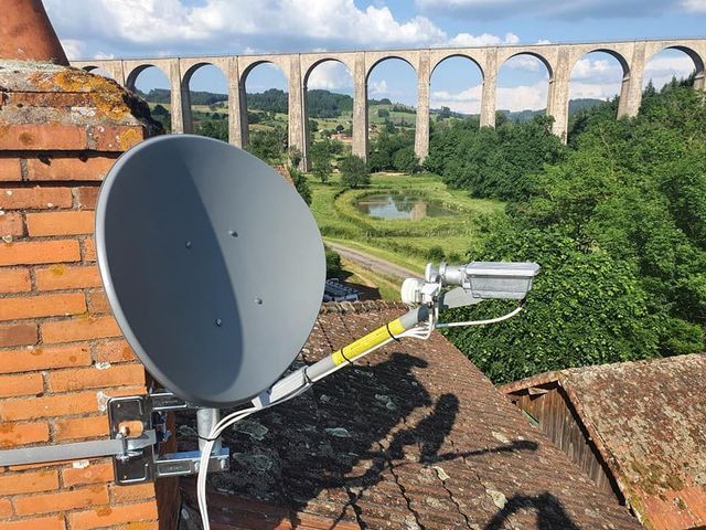 Antenne parabolique fixée sur une cheminée en briques, avec un grand pont en arc à l'arrière-plan. On aperçoit des arbres verdoyants et un ruisseau.