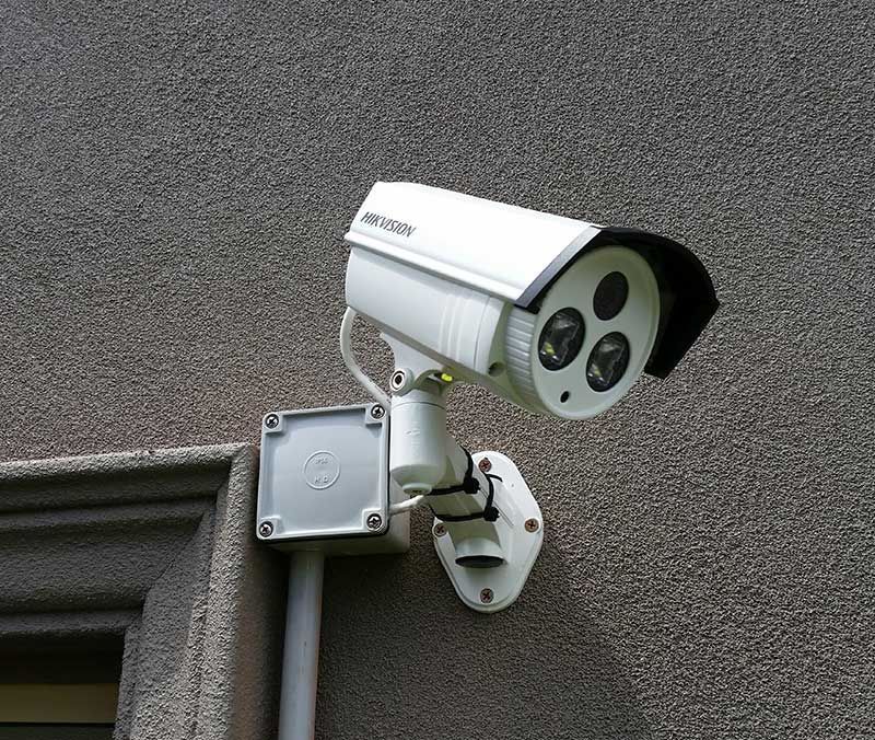 Caméra de sécurité blanche fixée sur un mur gris texturé, à côté d'un petit boîtier électrique blanc.