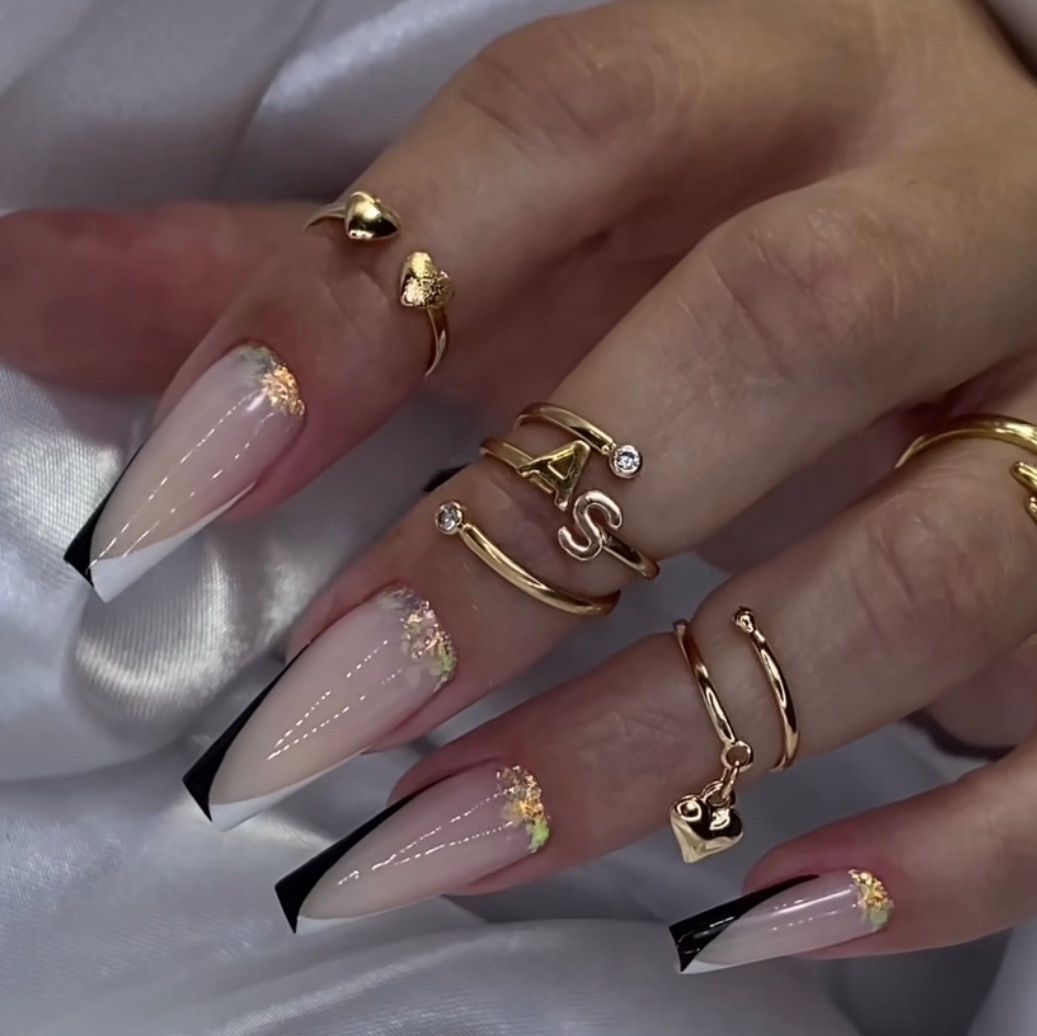 Primer plano de mano con uñas largas y elegantes en diseño negro y nude, adornada con anillos dorados.