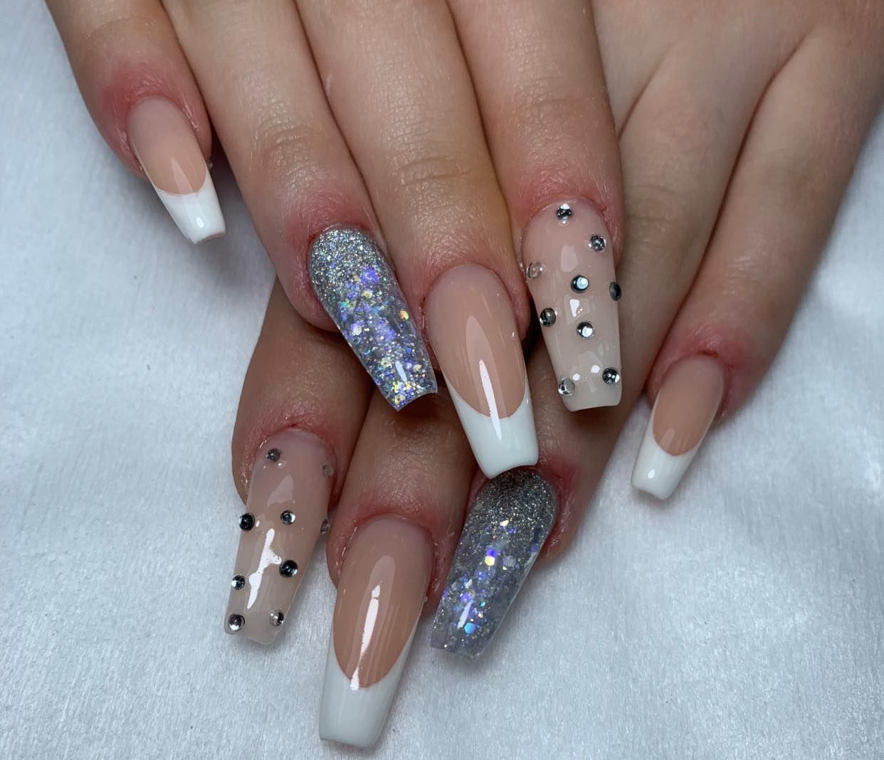 Una mano con uñas acrílicas largas y elegantes: puntas francesas, detalles de brillo y adornos de joyas.