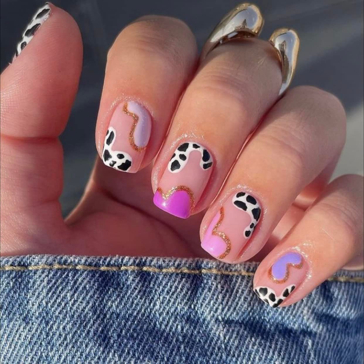 Mano con nail art: estampado de vaca y diseños en tonos rosa, morado y dorado sobre fondo azul jeans.
