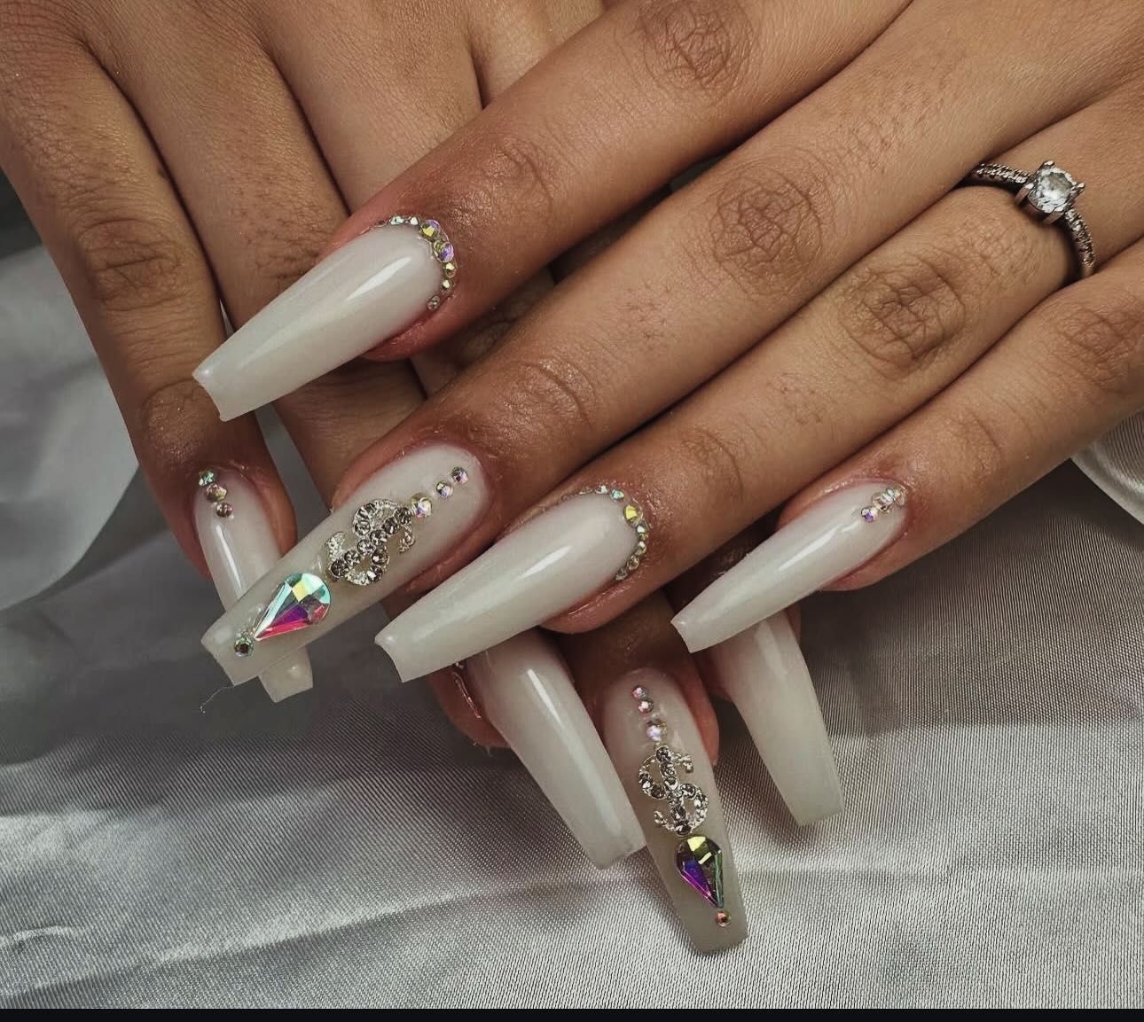 Uñas largas y blancas en forma de ataúd, decoradas con pedrería. Persona con un anillo de diamantes.