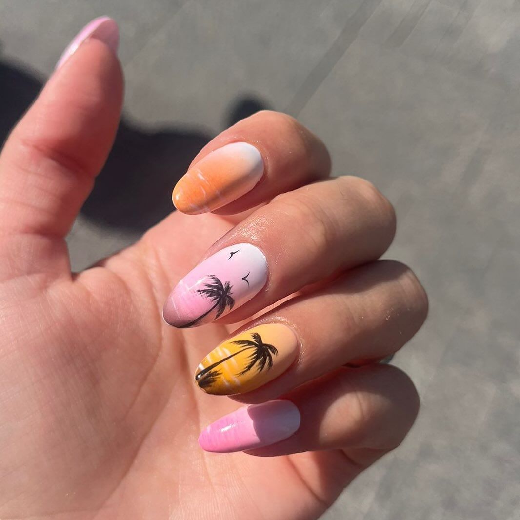 Mano con uñas ovaladas coloridas con palmeras y degradados de rosa, naranja y amarillo.