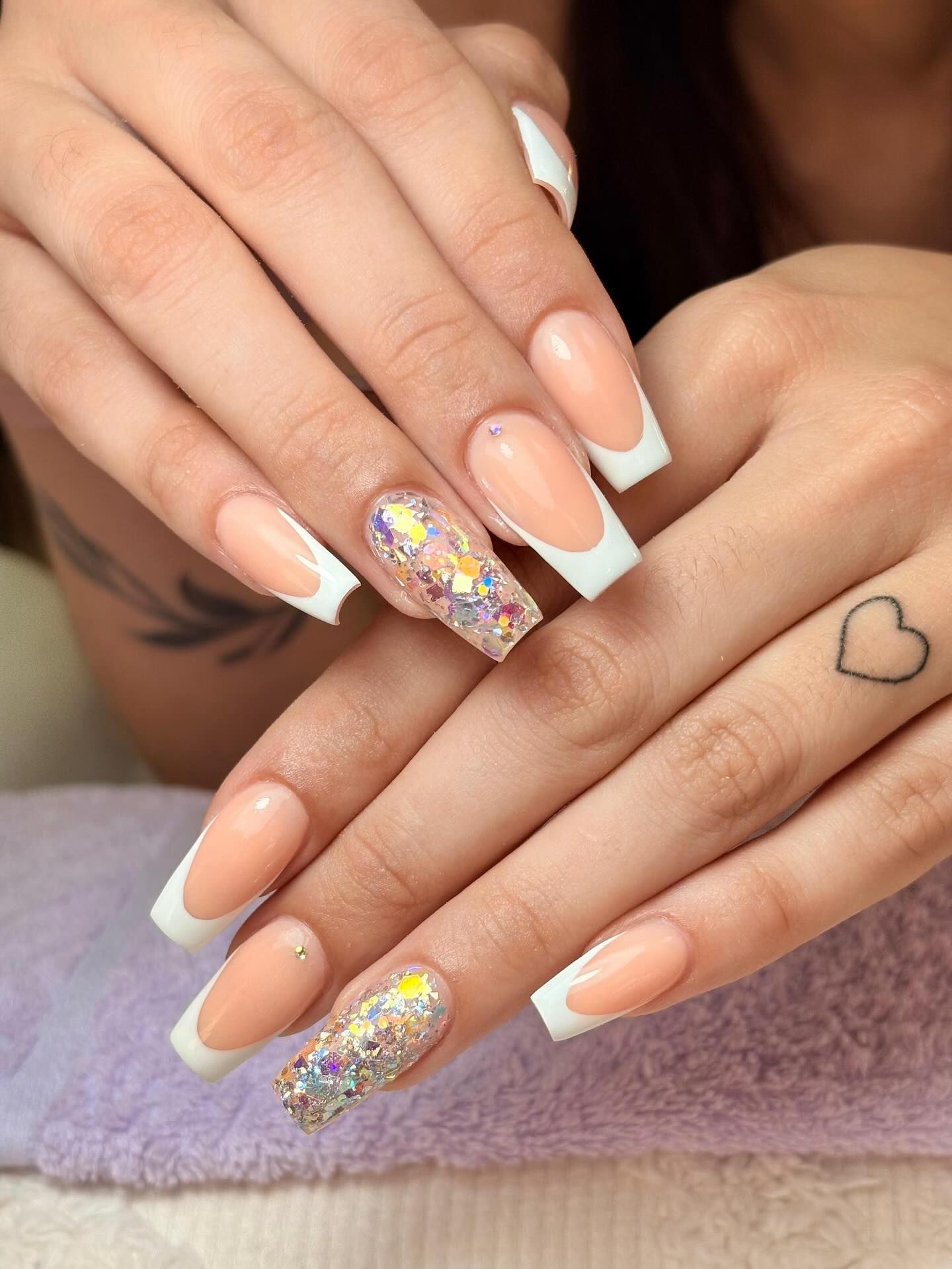 Manos con uñas francesas largas y cuadradas. Una uña tiene brillantina y un dedo tiene un tatuaje de corazón.