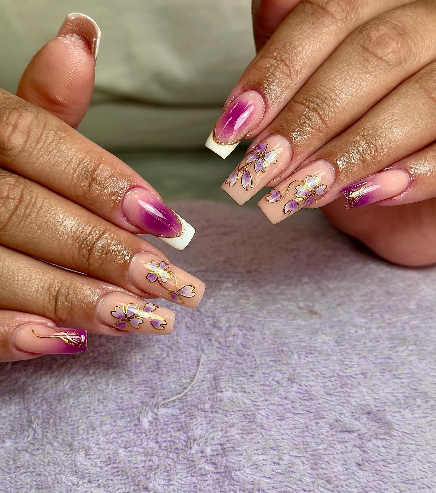 Uñas en degradado morado y nude con acentos florales dorados.