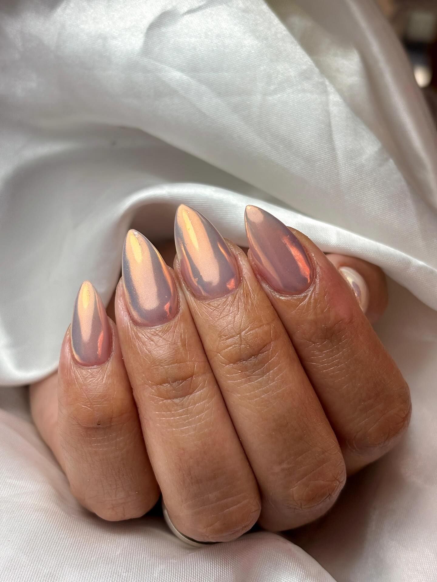 Mano con uñas esmaltadas en forma de almendra con un acabado brillante e iridiscente en tonos melocotón y morado.