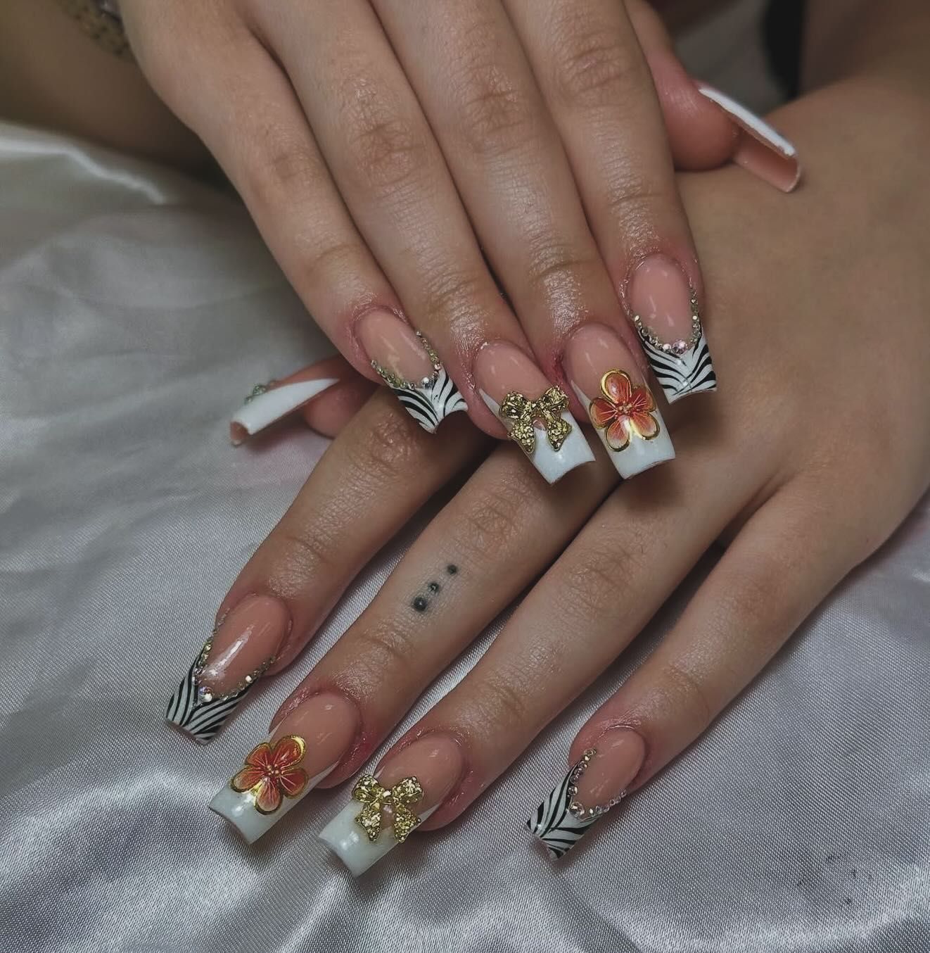 Manos con uñas acrílicas cuadradas, puntas francesas, estampado de cebra y detalles de lazos dorados.