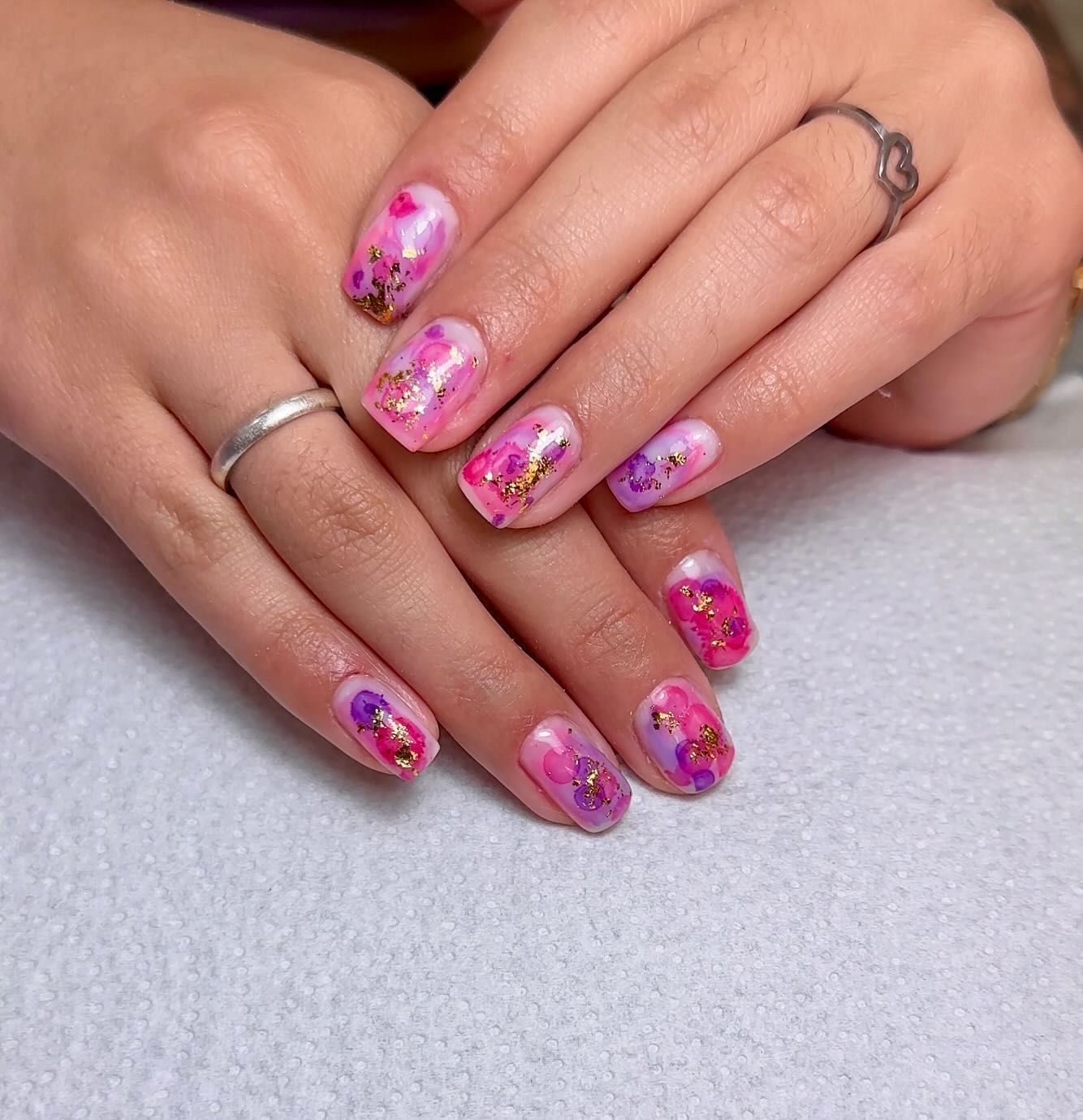 Arte de uñas floral rosa y morado con detalles de brillo dorado sobre un fondo claro.