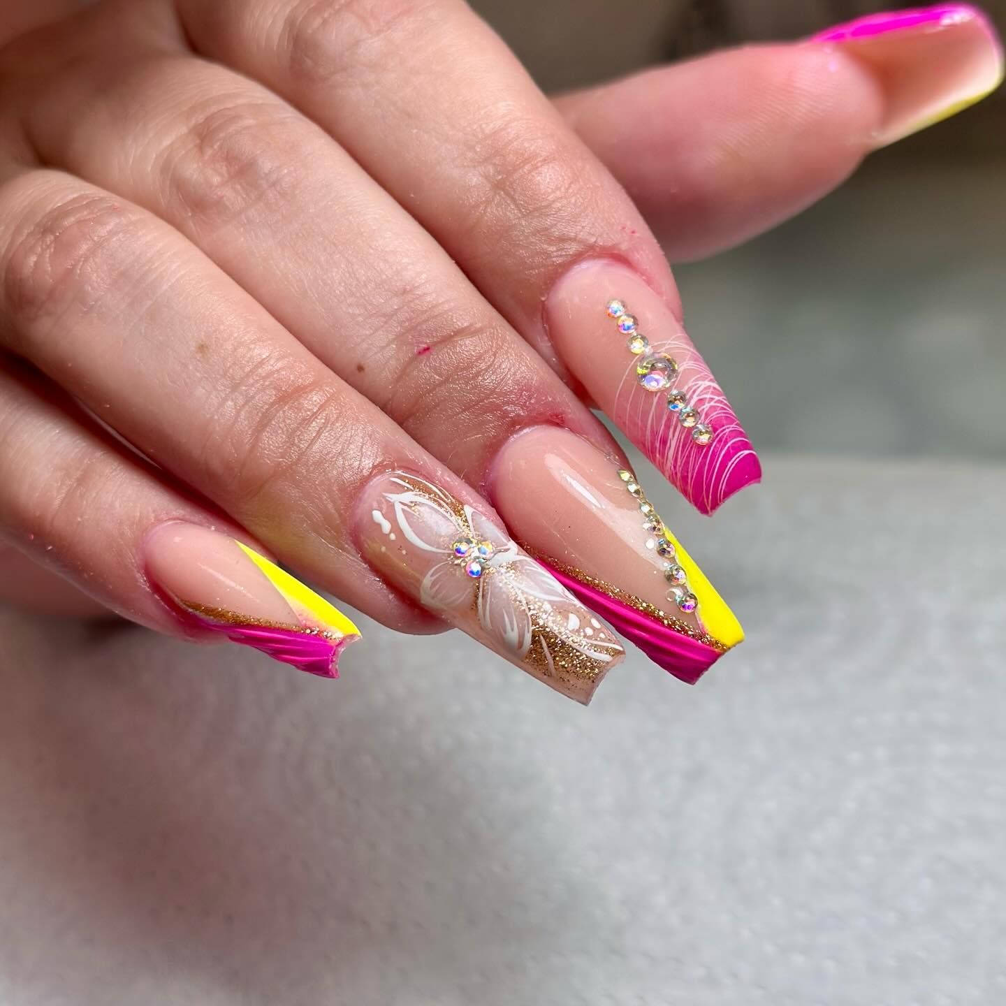 Primer plano de una mano con uñas acrílicas largas, cuadradas, de color rosa, amarillo y transparente, decoradas con diamantes de imitación.