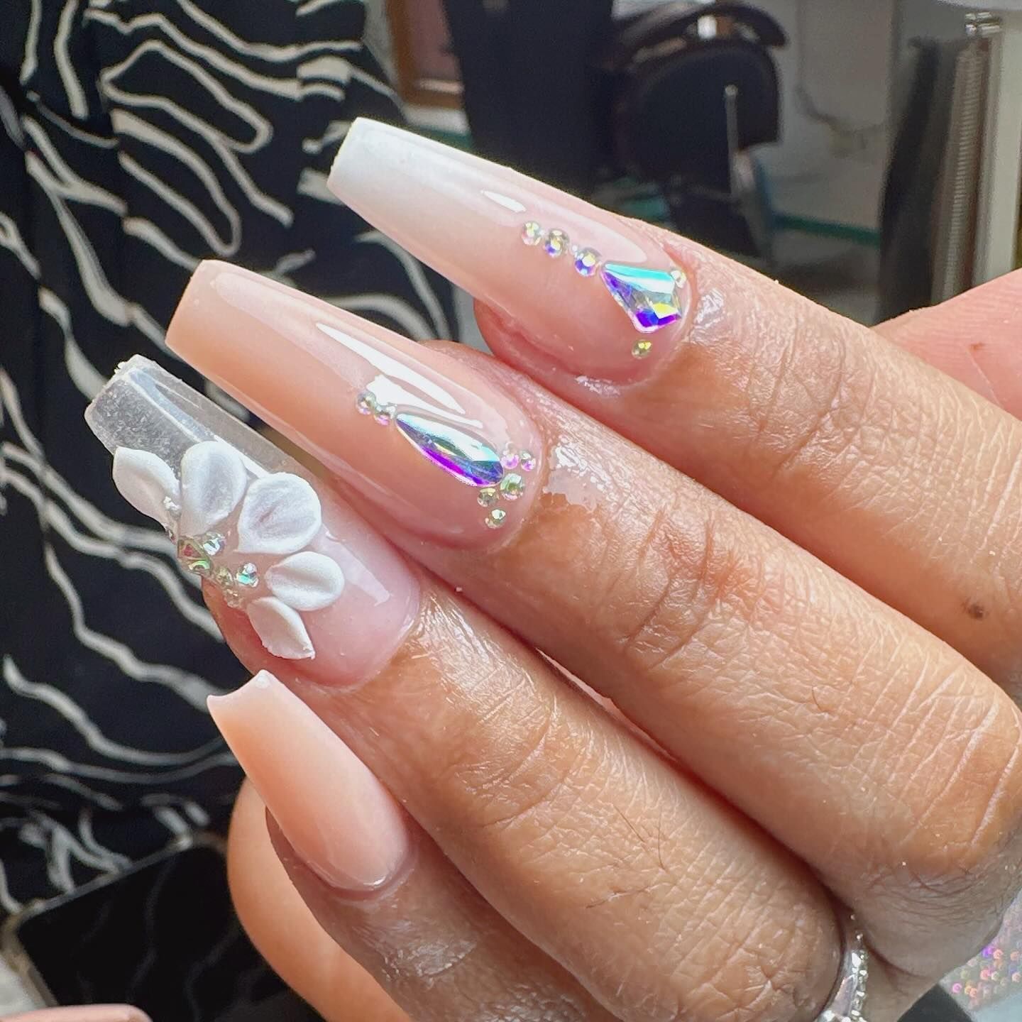 Uñas acrílicas largas en forma de ataúd en color nude, con ombré blanco, pedrería y detalles de flores blancas en 3D.