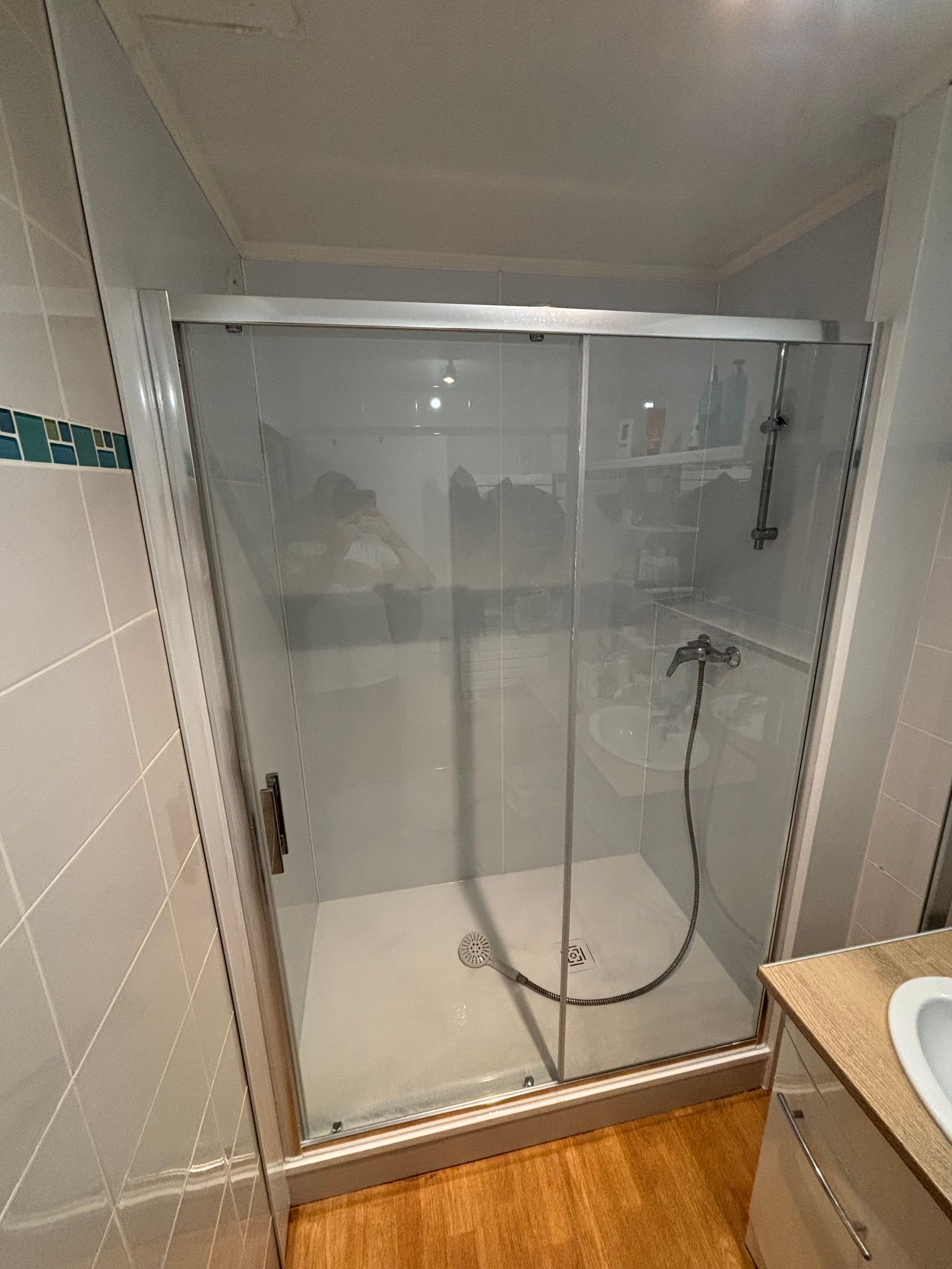 Petite cabine de douche avec portes en verre, robinetterie chromée et sol imitation bois.