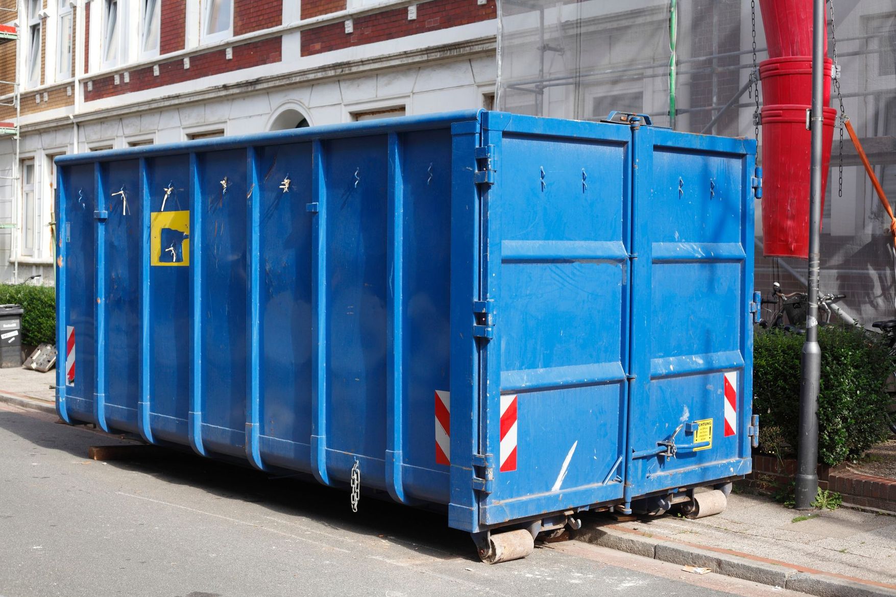 Une grande benne à ordures industrielle bleue est garée dans la rue, à côté d'un bâtiment en briques et d'une clôture.