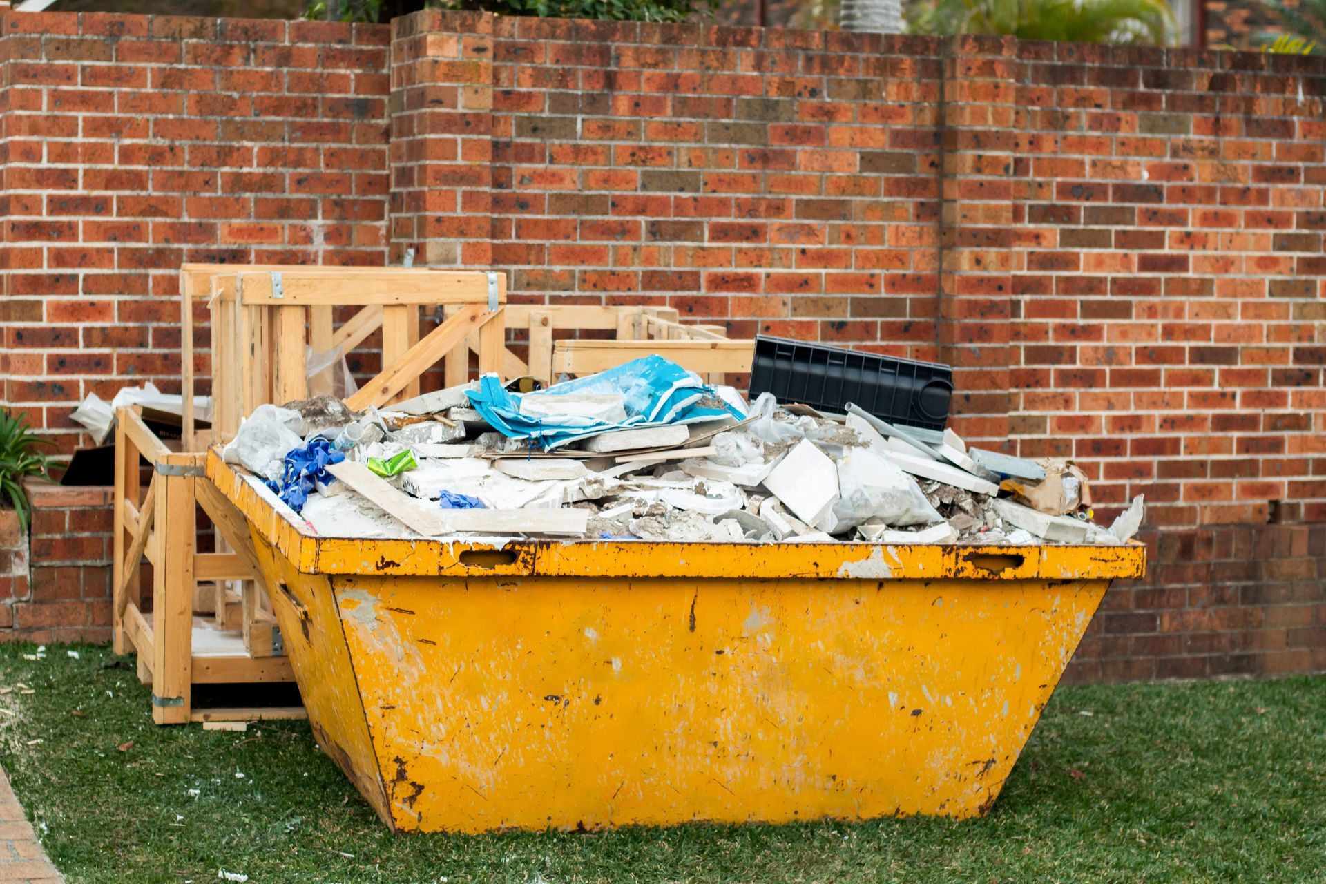 Un conteneur à déchets jaune rempli de débris est adossé à un mur de briques et à une palette en bois à l'extérieur.