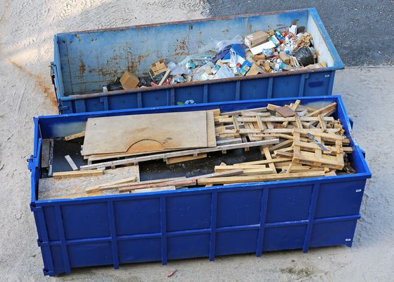 Deux bennes bleues remplies de débris de construction et de chutes de bois sur le trottoir.