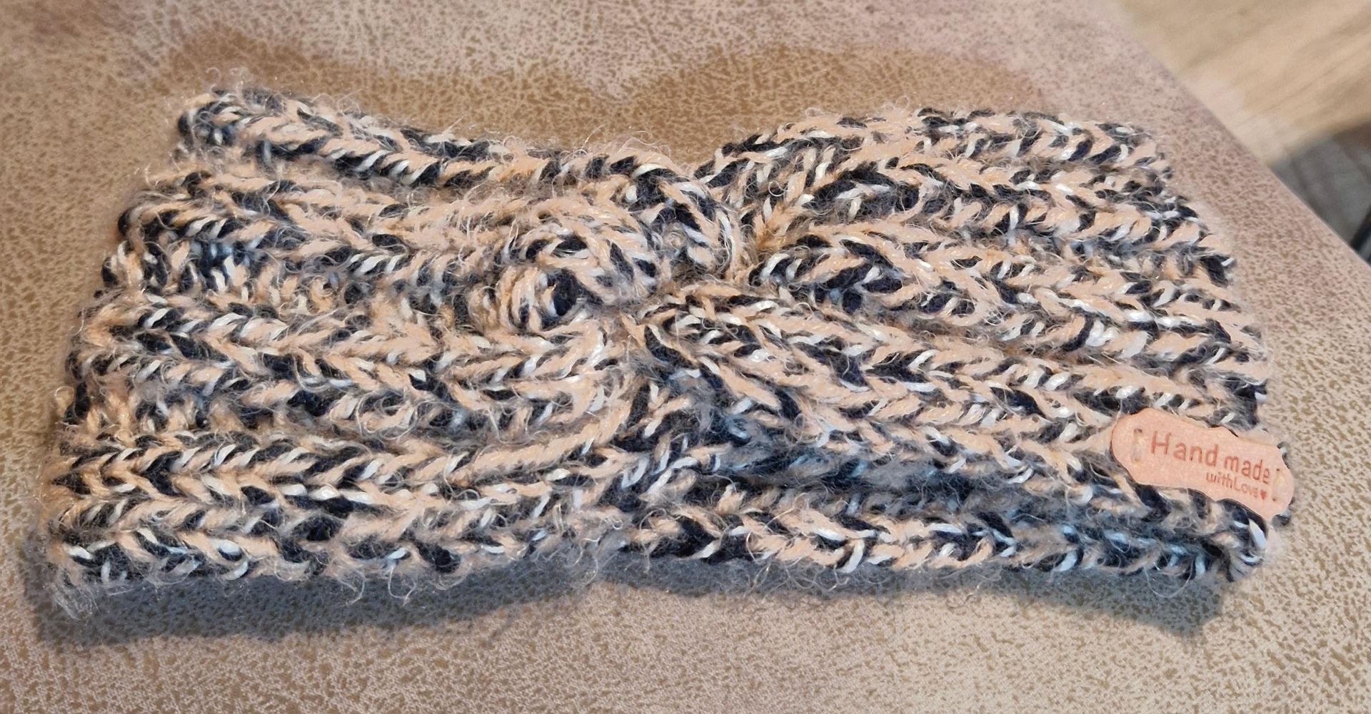 Un bandeau tricoté torsadé en fil marbré beige, crème et bleu foncé, avec une petite étiquette « Fait main » sur le côté.