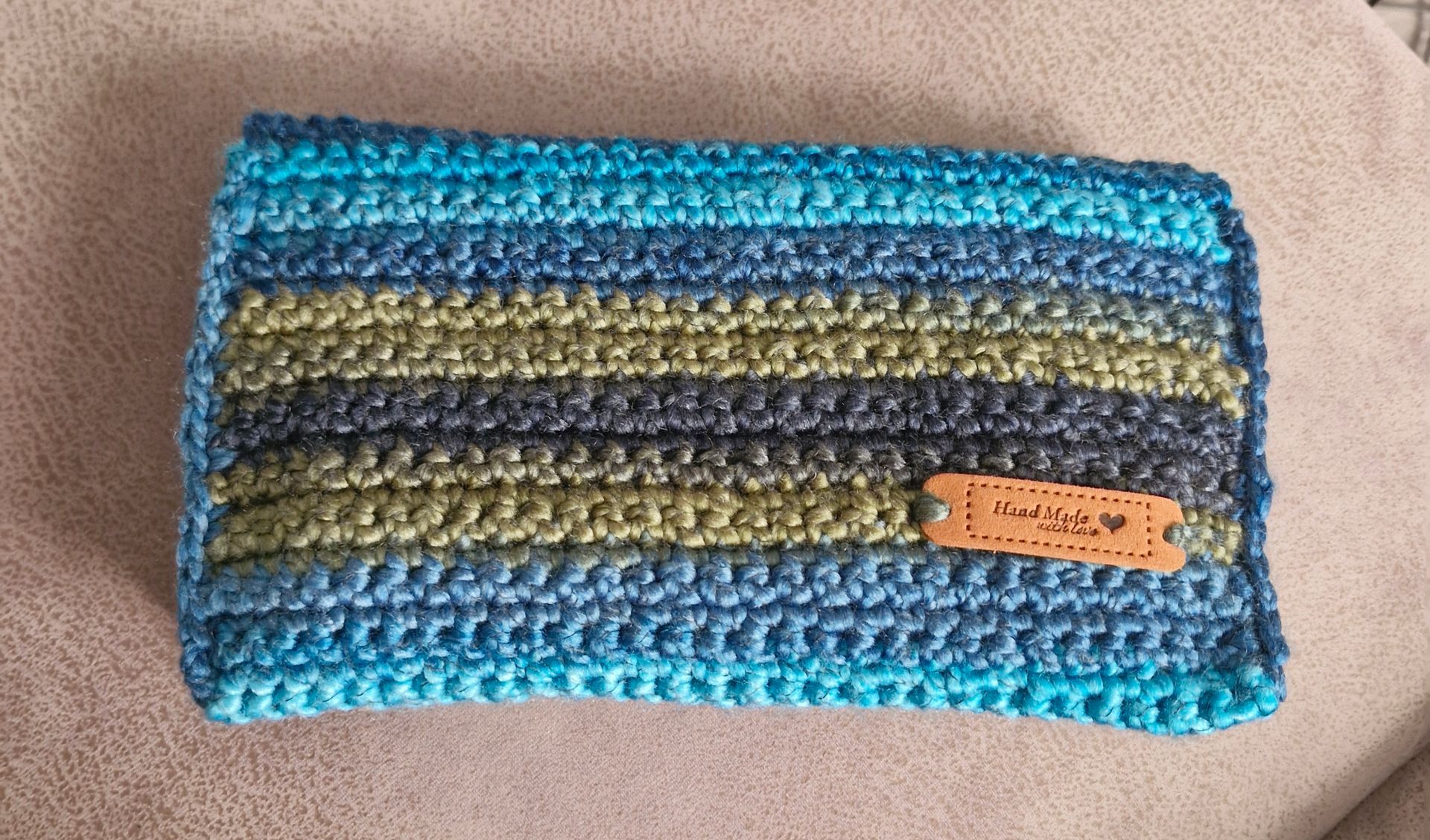 Une pochette rectangulaire au crochet faite main, ornée de rayures horizontales en fil bleu, vert et gris, avec une étiquette en cuir.