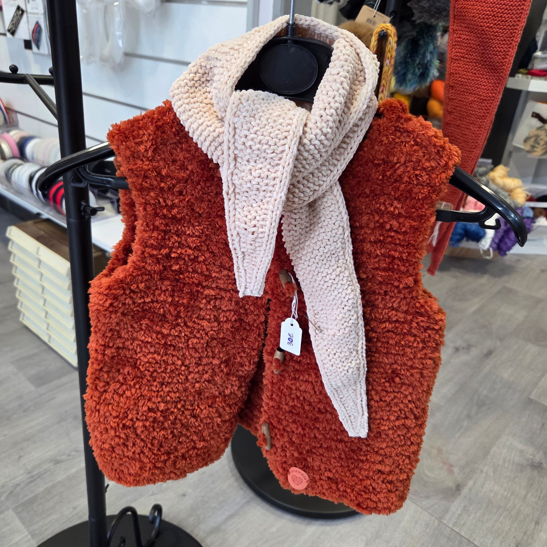 Un gilet boutonné duveteux, de couleur orange rouille, associé à une écharpe en tricot couleur crème, est suspendu à un présentoir dans un magasin de loisirs créatifs.