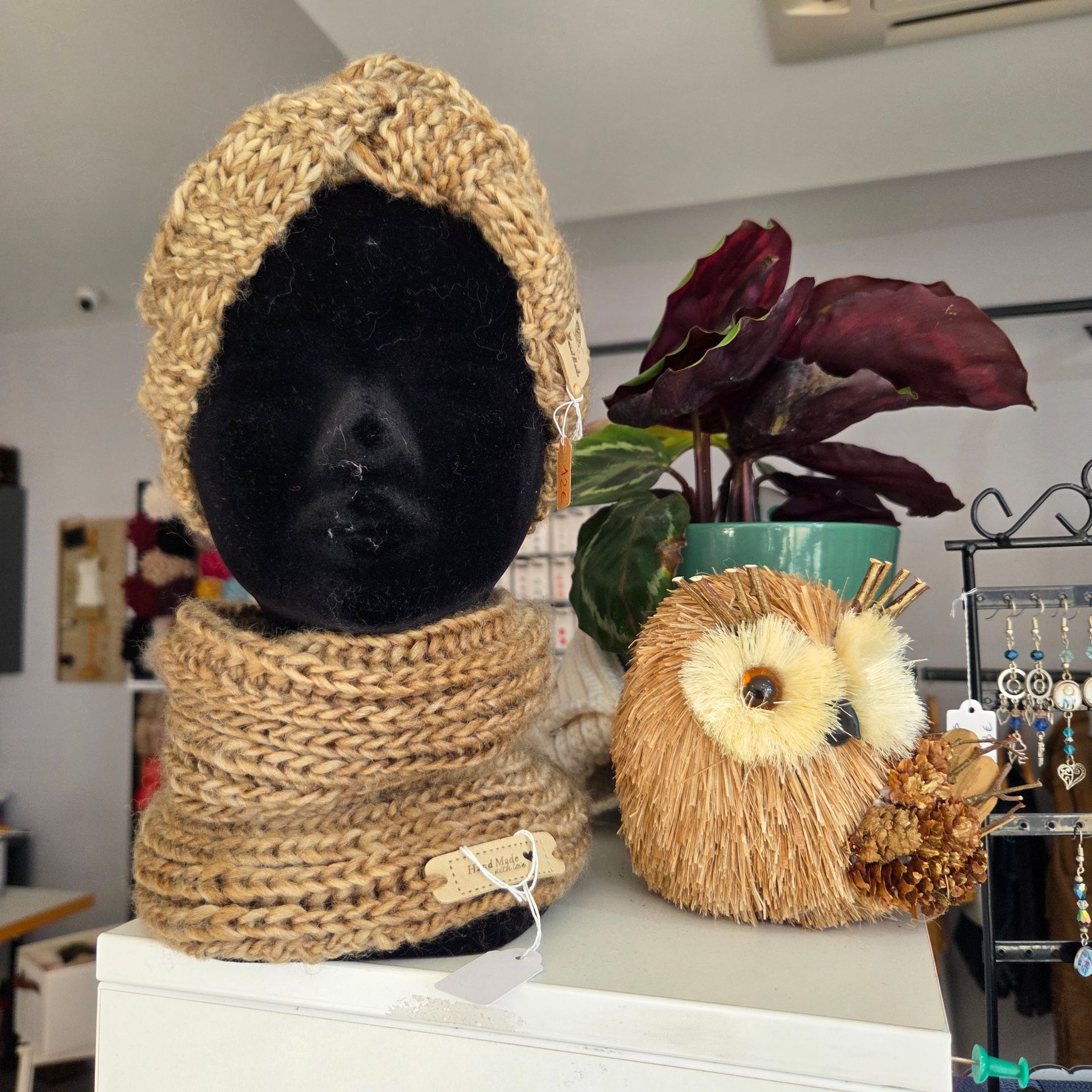 Une tête de mannequin noire portant un ensemble bandeau et écharpe en grosse maille beige, à côté d'une chouette décorative en paille et d'une plante.