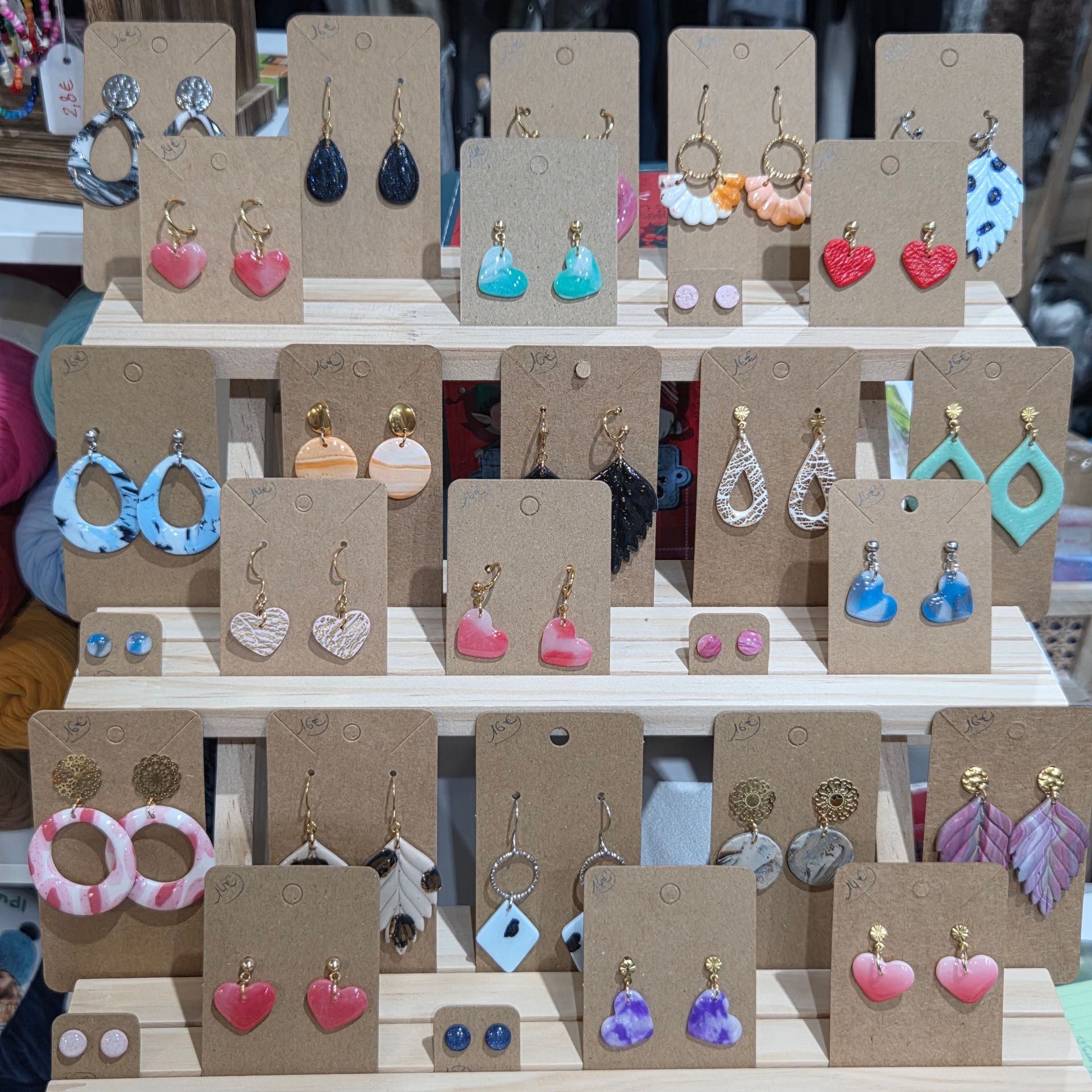 Un présentoir contenant des dizaines de paires de boucles d'oreilles pendantes et à clous colorées, faites à la main, sur des supports individuels en carton.