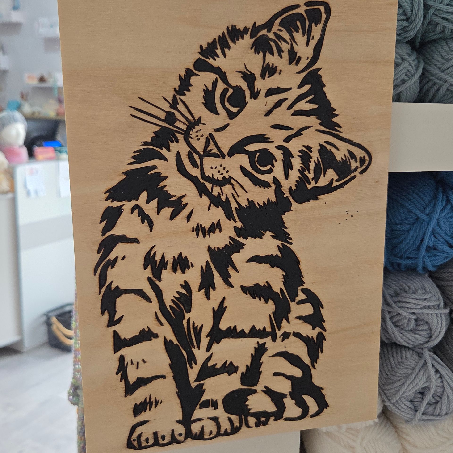 Un dessin au trait noir représentant un chaton tigré la tête inclinée, imprimé sur une planche de bois rectangulaire verticale.